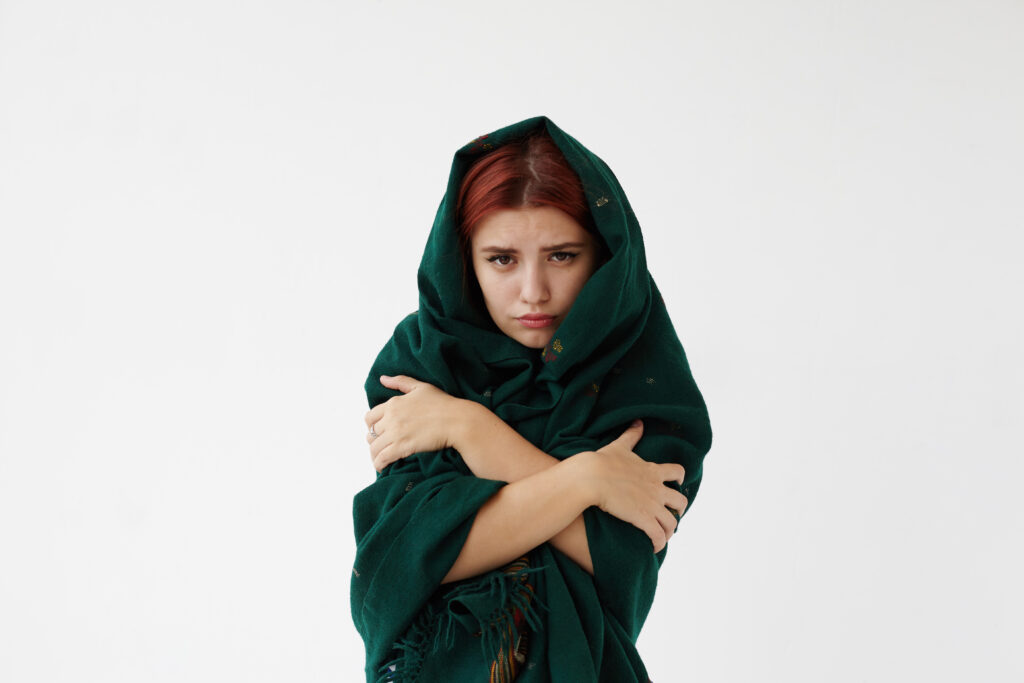 Horizontal shot of cute young female wrapping herself in warm dark green plaid, feeling cold because of high temperature, looking at camera with sad and happy expression. People and sickness